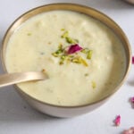 A metal bowl of creamy rabdi garnished with chopped pistachios and a rose petals.