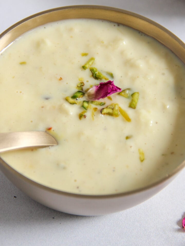 A metal bowl of creamy rabdi garnished with chopped pistachios and a rose petals.