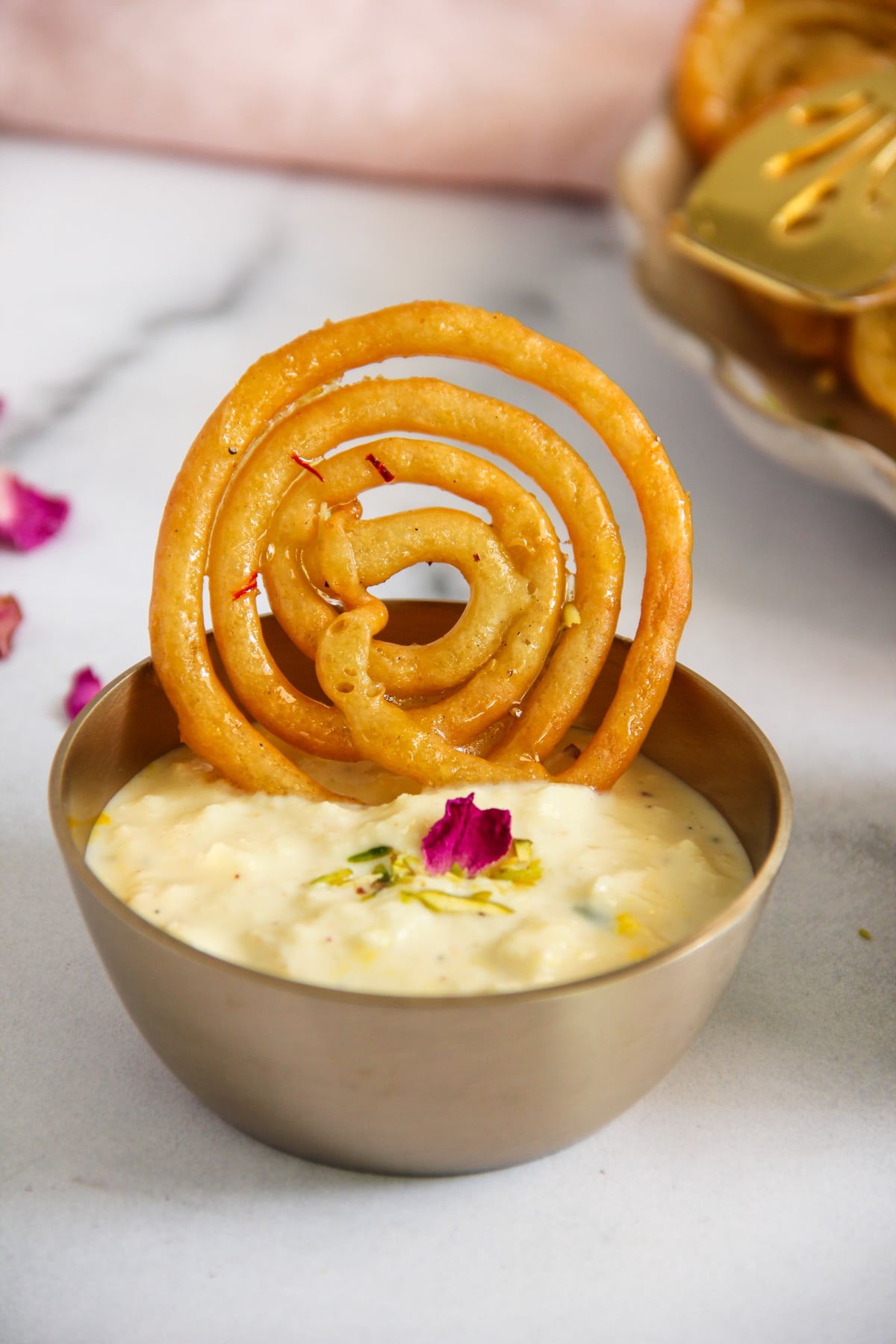 A spiral jalebi resting in a bowl of creamy rabri topped with rose petals and pistachios.