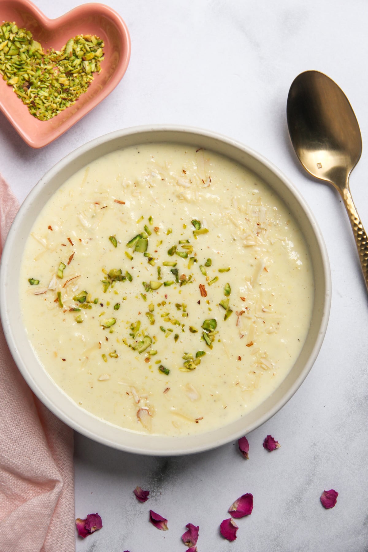 A bowl of creamy rabdi garnished with chopped pistachios, next to a small heart-shaped dish of pistachios and a spoon.