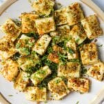 A plate of cubed, pan-seared paneer cheese garnished with chopped herbs and spices.