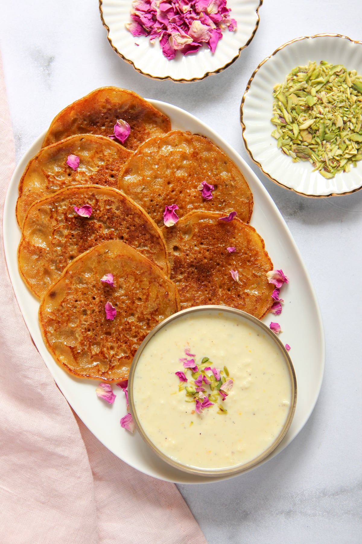A plate of golden-brown malpua sprinkled with dried rose petals, served with a bowl of rabdi, chopped pistachios, and additional rose petals.