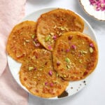 Four golden-brown malpuas on a white plate, garnished with chopped pistachios and dried rose petals.