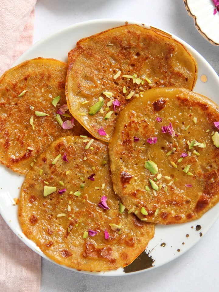 Four golden-brown malpuas on a white plate, garnished with chopped pistachios and dried rose petals.