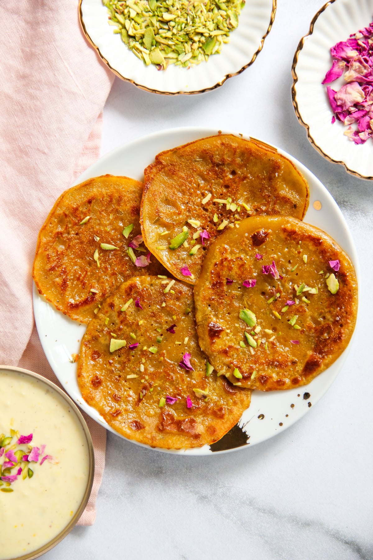 A white plate topped with four golden-brown malpuas garnished with pistachios and rose petals, served with a bowl of rabdi.