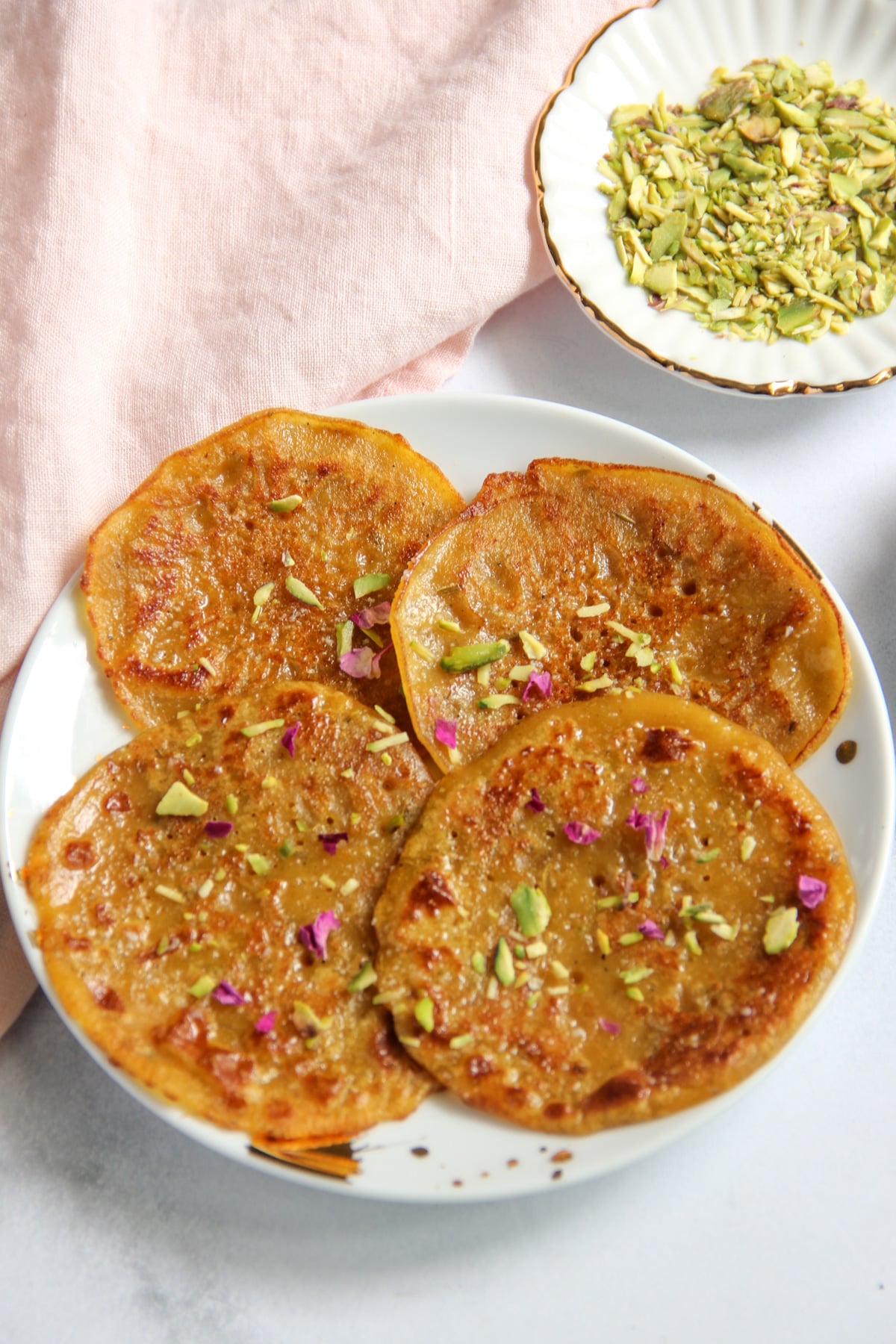 Four golden, syrup-soaked pancakes on a plate, garnished with chopped pistachios and dried rose petals, with a small bowl of crushed pistachios nearby.