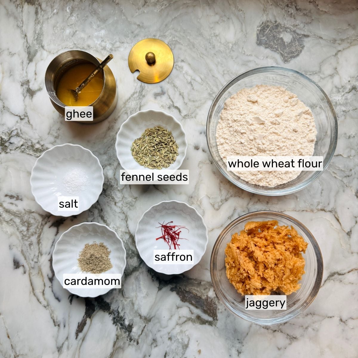 Malpua ingredients on a marble surface: ghee, fennel seeds, whole wheat flour, salt, cardamom, saffron, and jaggery, each labeled with its name.