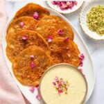 Oval platter of pan-fried malpua garnished with rose petals, served with a bowl of rabri, alongside small dishes of chopped pistachios and rose petals.