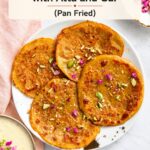 A plate of four golden-brown malpua pancakes topped with chopped pistachios and rose petals, with text overlay reading Easy Malpua Recipe with Atta and Gur (Pan Fried).